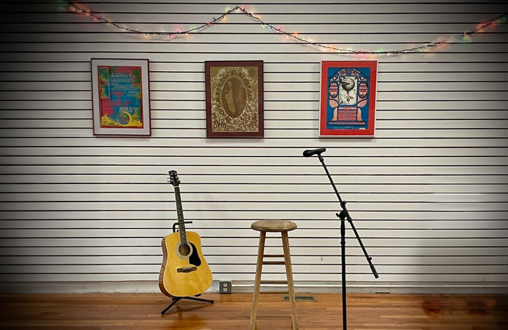 A guitar on a stand, a stool and a microphone stand sit in front of a white slatted wall with art hanging on it. A string of lights are strung above the artwork on the wall