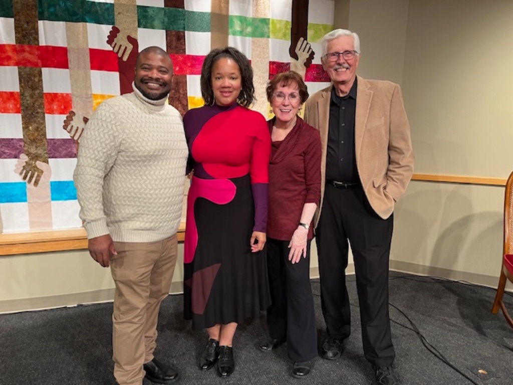 A black man in a white sweater and khaki pants and a black woman in a red, purple, and black dress a white woman in a maroon shirt, and a white man with white hair in a black shirt and pants with a tan jacket stand in front of a plaid quilt made up of crossed arms holding hands.