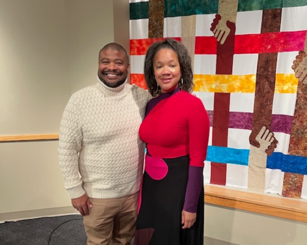 A black man in a white sweater and khaki pants and a black woman in a red, purple, and black dress stand in front of a plaid quilt made up of crossed arms holding hands.