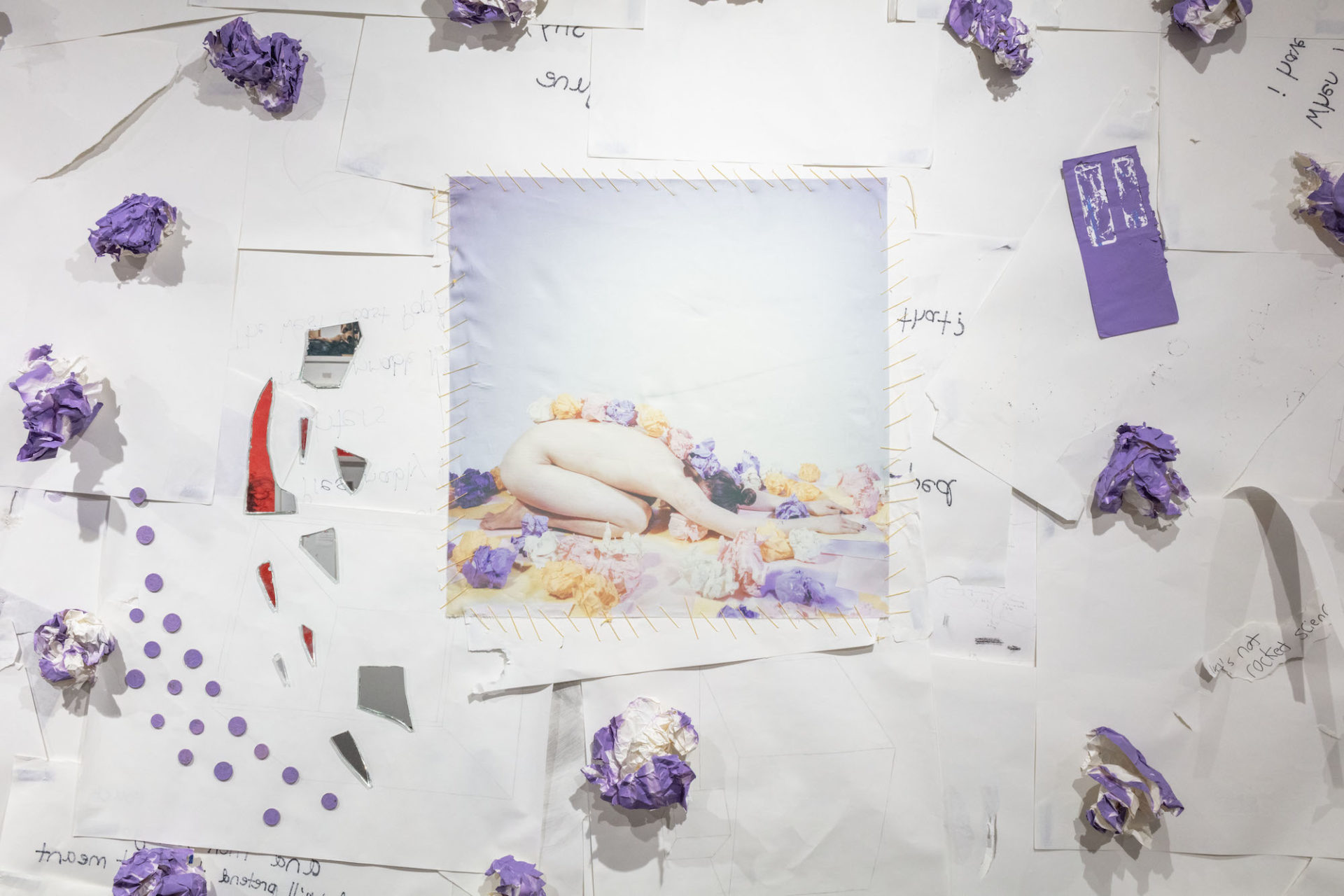 Multimedia artwork with a white paper overlapping to create the background with purple three dimensional pieces scattered about. In the center is a square photograph sewn onto the background. The photo is of a naked person in child's pose surrounded by more of the same purple crumpled papers including a line of them down the person's back as they are on the ground with their face to the ground and arms extended in front.