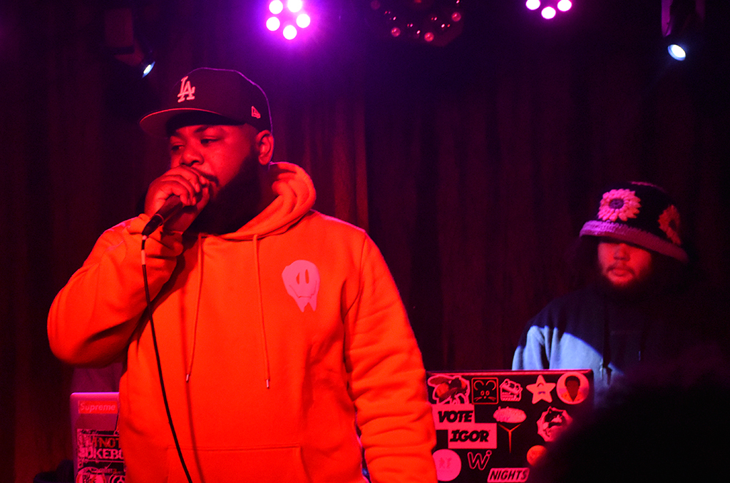 Two individuals are on a stage set against a backdrop of a red curtain. The individual on the left is attired in a yellow hoodie featuring a white alien head design, complemented by a black baseball cap emblazoned with a white “LA”. The individual on the right is dressed in a blue hoodie and also sports a black baseball cap with a white “LA”. The stage is bathed in a mix of pink and purple lights, adding to the ambiance. A laptop adorned with stickers, is visible in the background.