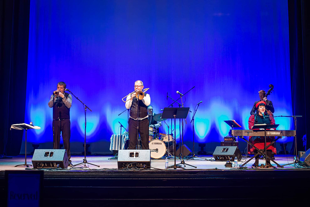 The scene captures a moment on a stage where four musicians are immersed in their performance. Each musician is attired in a black suit and hat, adding a touch of formality to the event. The stage is bathed in a soft blue light, creating a serene ambiance. In front of the musicians, music stands and microphones are set up, ready to capture every note. The backdrop is a simple blue curtain, keeping the focus on the performers.