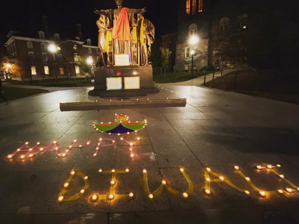 in the foreground candles spell out Happy Diwali, behind it is a design made out of different colors of sane and the Alma Matter lit up in orange in the background. 