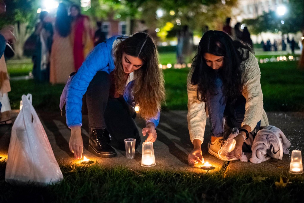 Two young women with long dark hair kneel down on the sidewalk to light small candles.