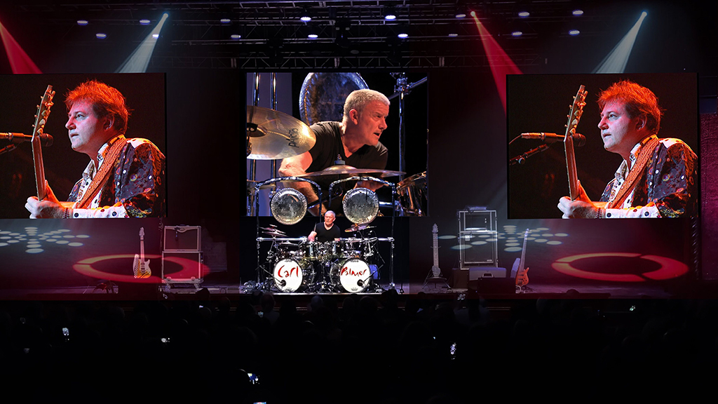 The picture captures a stage scene illuminated by red and white lights. Three large screens display a drummer in a black shirt and two guitarists in white jackets with sequins, all engaged in a performance. The atmosphere suggests a live concert.