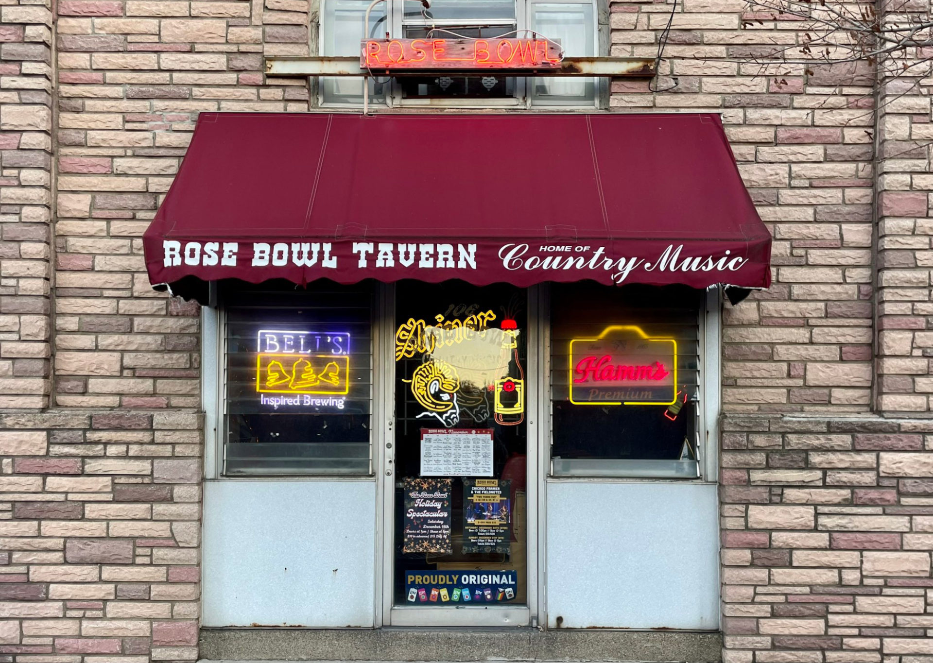 Old entrance to Rose Bowl Tavern. An unusable door / entry in the middle of a blonde brick building. There is a maroon awning over the door that says "Rose Bowl Tavern." There are some neon lights for beer in the window.