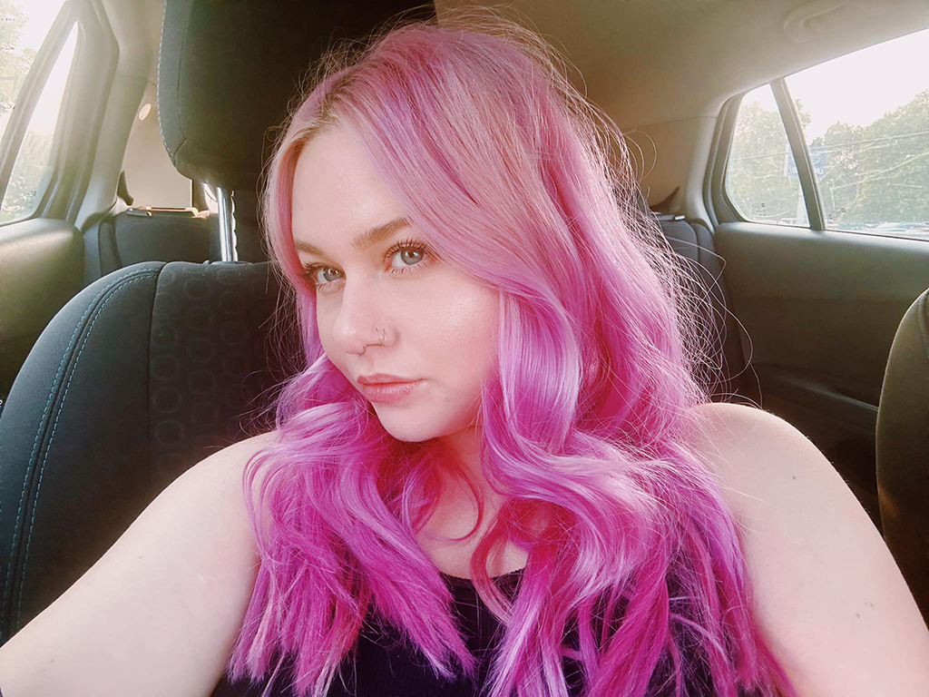 A girl with long pink hair in the passenger seat of a car looking out the window.