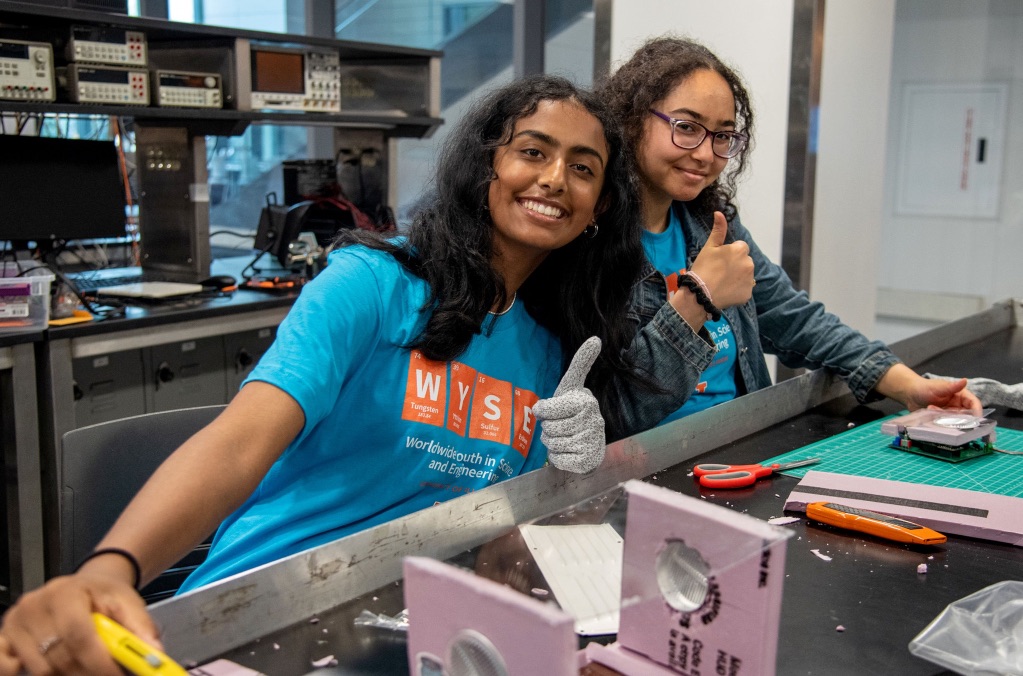 Two brown woman in matching bright blue shirts sit at a big table in front of machinery. They both have long dark hair and one is giving a thumbs up to the camera.