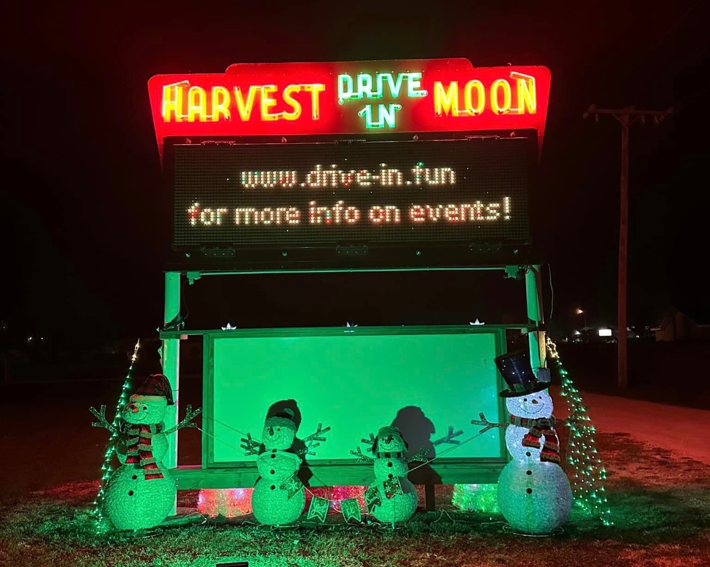 The Harvest moon drive-in sign is illuminated in red and green neon letters with snowman standing in front of a bright green light with their stick arms in the air. 