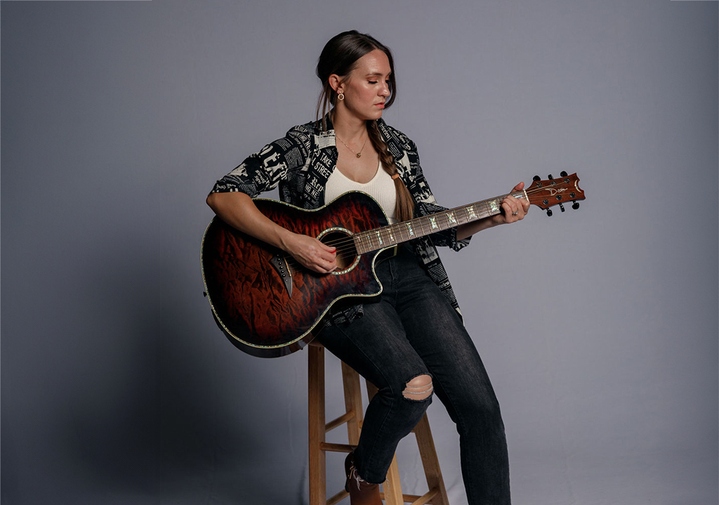 A woman with black hair sits on a stool holding a guitar against a grey background.
