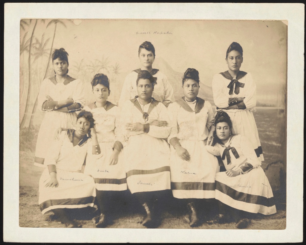 A sepia toned photograph of a group of woman who were members of the Royal Hula troupe. They are all wearing matching long white dresses with a dark stripe on the bottom.