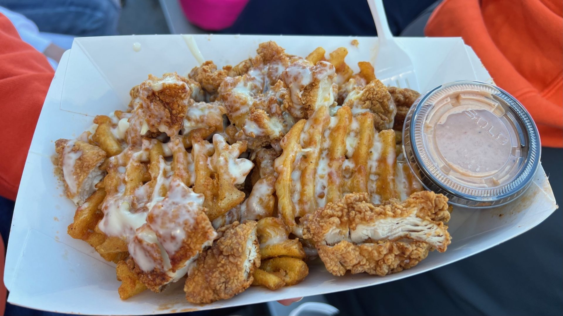 A rectanguler paper tray filled with waffle fries, chunks of breaded chicken, and covered in a white sauce.