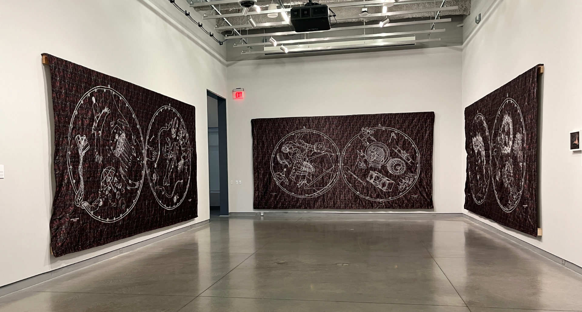 Three huge purple-ish tapestries with white embroidery on them are on three walls in a gallery. Each tapestry contains two circles with imagery inside; they resemble star maps.
