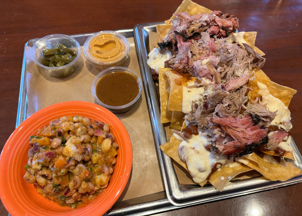 On the right: A plate of pulled pork nachos at Black Dog. Golden chips are piled with white cheese and pulled pork on a rectangular silver tray.  On the left: three small containers with pickled jalapeños, BBQ ranch, BBQ sauce, and an orange bowl of Texas caviar. 