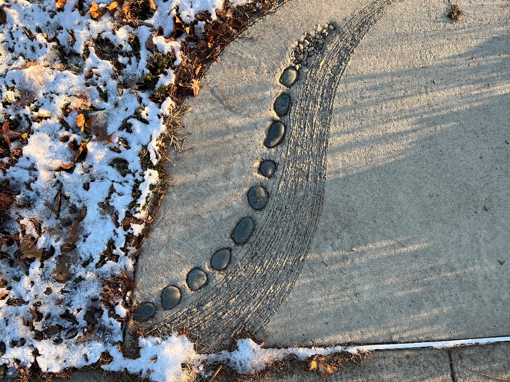 A curved sidewalk, inlaid with rocks and a raked wave pattern.. On the left edge are brown leaves and a dusting of white snow. 