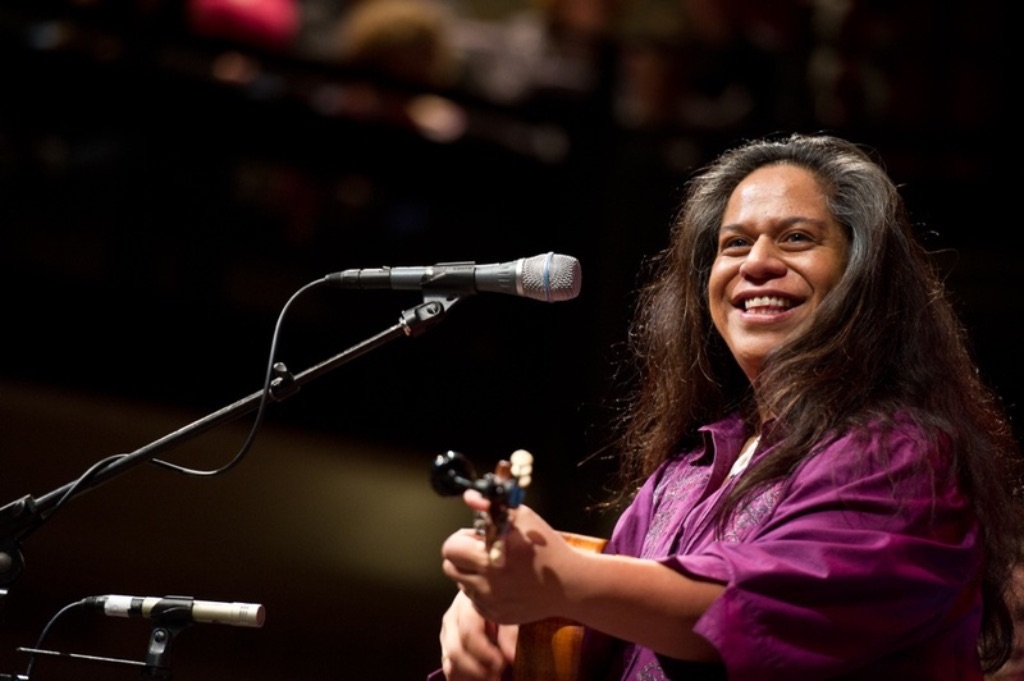 A brown woman with long dark hair stands in front of a microphone stand and  plays a ukulele. She is wearing a purple collared shirt. 