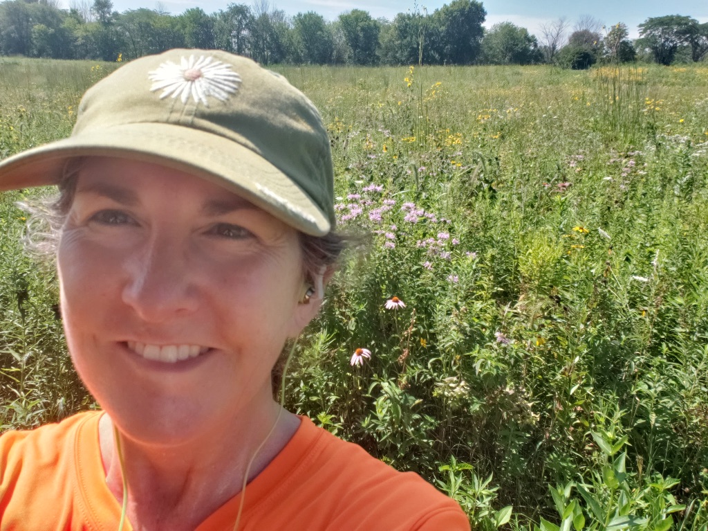 A white woman in a beige hat with a white embroidered daisy on it and an orange t-shirt stands in front of a field of flowers on a sunny day. 