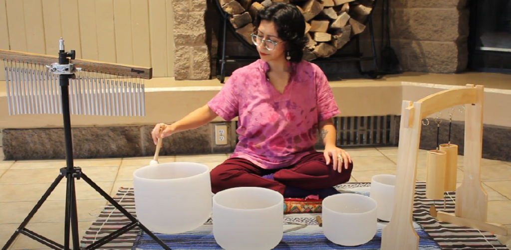 A woman with dark hair and glasses sits cross-legged on a rug. She has an array of sound bowls and chimes placed around her.