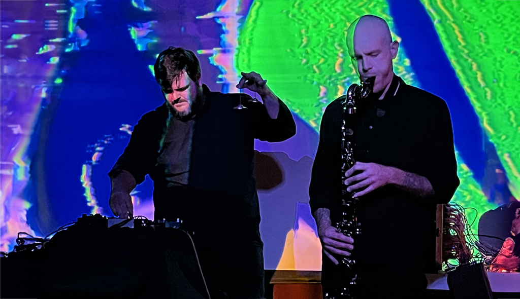 Two individuals are standing on a stage with a vibrant background of blue, green, and yellow hues. The individual on the left is attired in a black shirt and is seen holding a microphone. On the right, the individual is dressed in a black suit and is seen holding a saxophone.