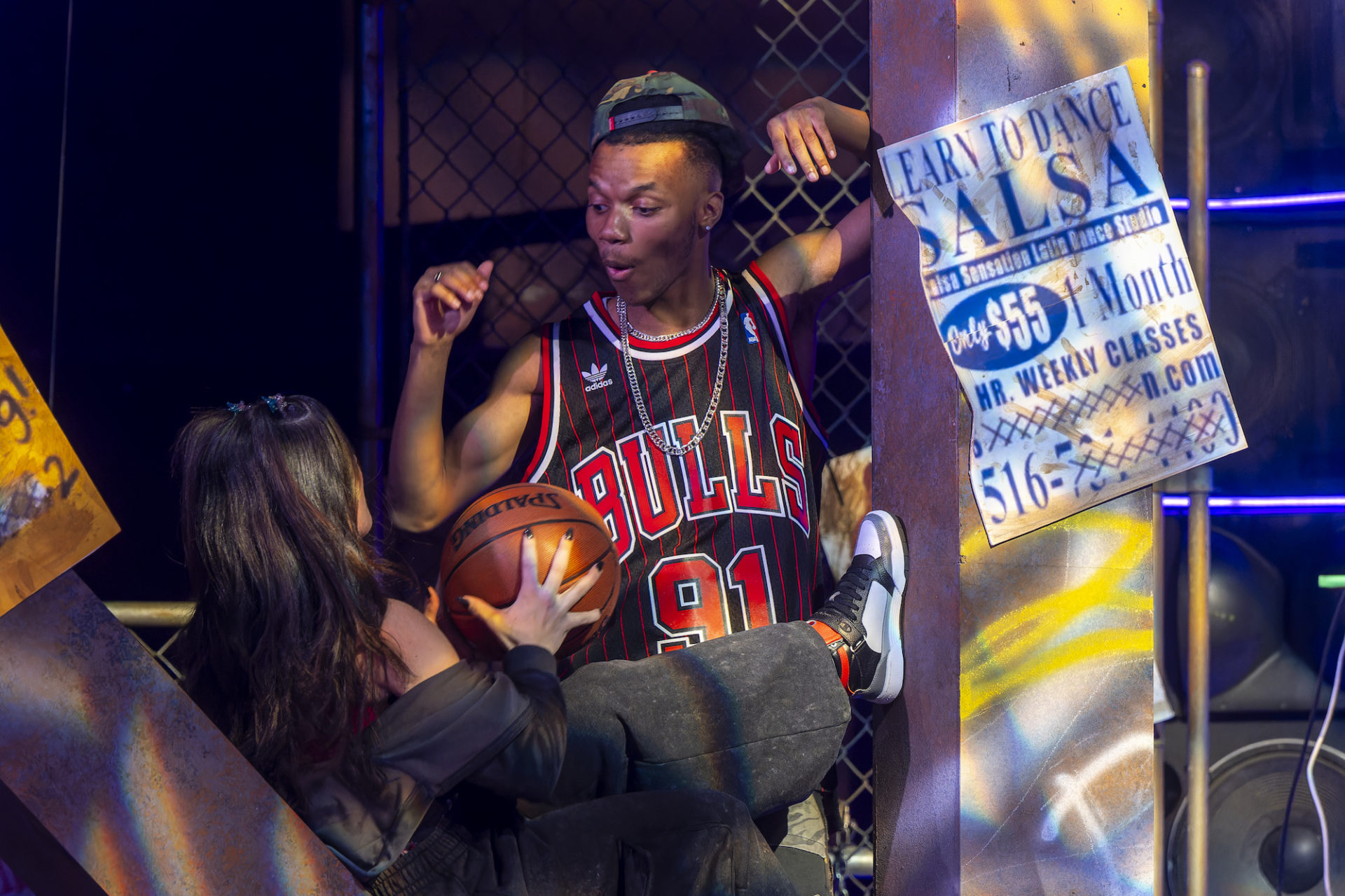 A scene from the Realness: a stage with exposed brick and posters hanging; a Black man in a Bulls jersey looks surprised while a woman holds a basketball
