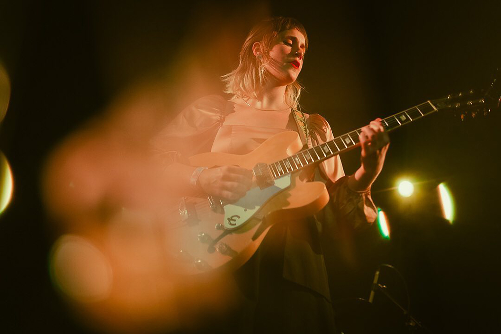 A person is on a stage, playing an electric guitar. They are dressed in a white top and have blonde hair. The guitar is white. The stage is lit with lights in the background.

