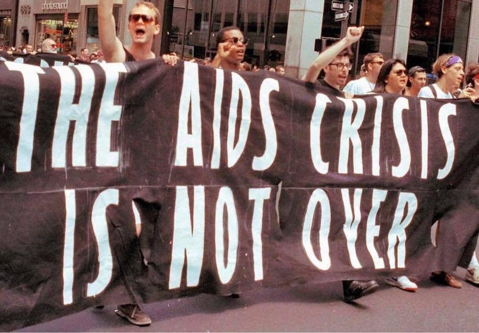A line of people holding a large black banner with white block letters that say The AIDS Crisis is Not Over.