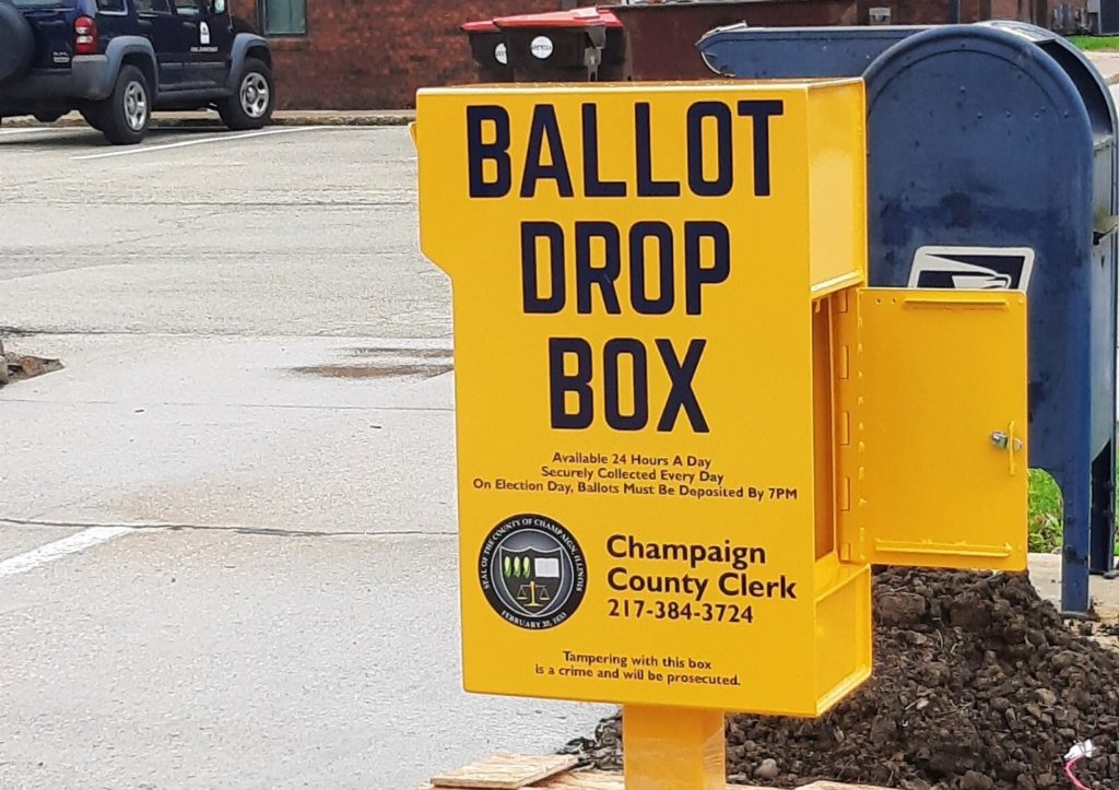 A yellow metal box on a stand next to a parking lot. It says Ballot Drop Box in blue block letters.