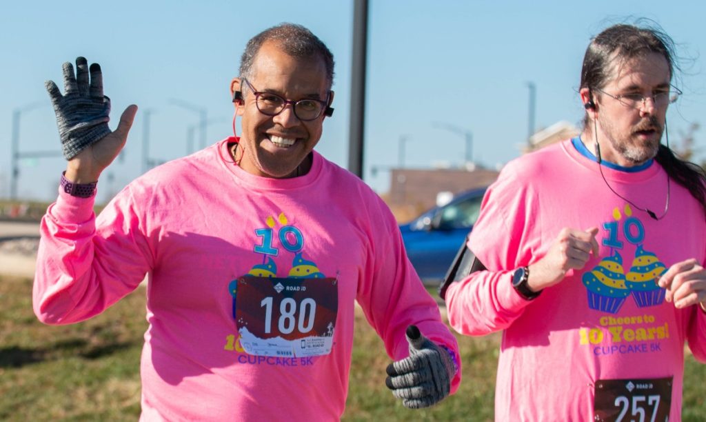 A Black man with short hair and glasses and a white man with long hair and glasses are both wearing pink long sleeve t-shirts and running alongside each other.