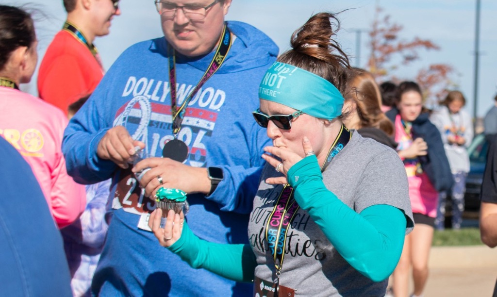 a white woman in a teal headband and long sleeve shirt with a gray shirt over it licks her finger and holds a chocolate cupcake with teal icing in the other hand. She is surrounded by other runners. 