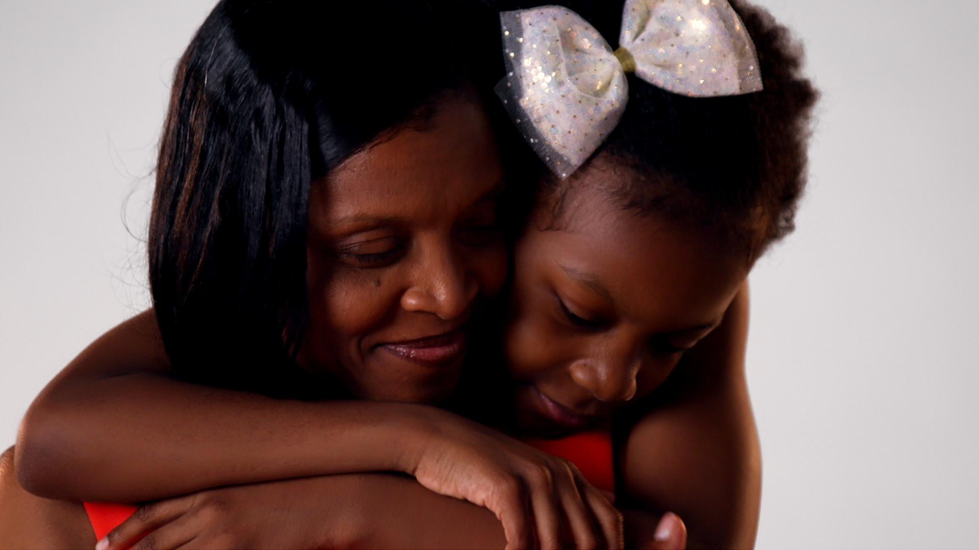 A young Black girl is hugging a Black woman from behind. They are both smiling.