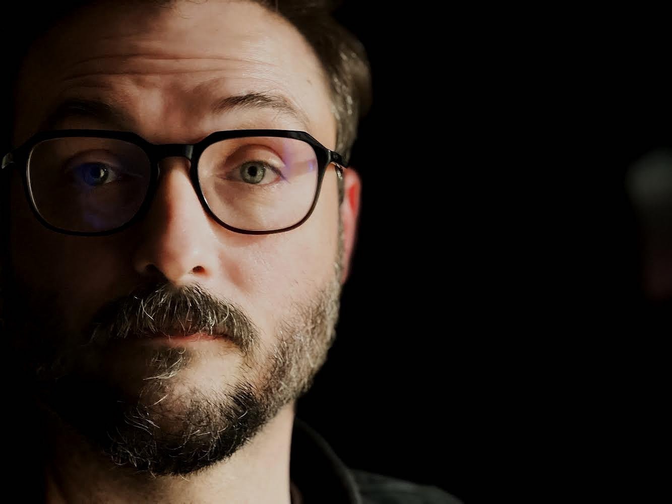 Caleb Curtiss, a white man with a beard and black glasses, stares at the camera, unsmiling. The photo is a close-up so that only his face is visible against a solid black background