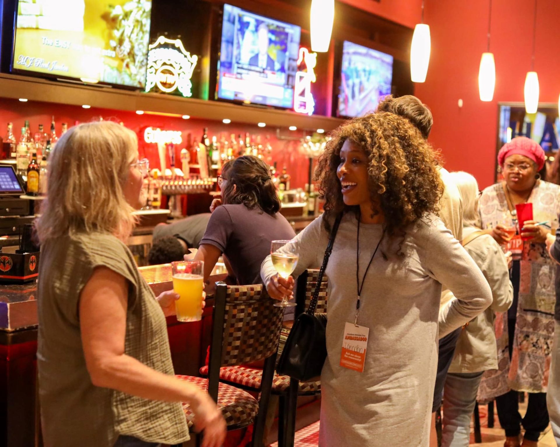 A Black woman holding a glass of wine and a white woman holding a beer and conversing near a bar filled with people.
