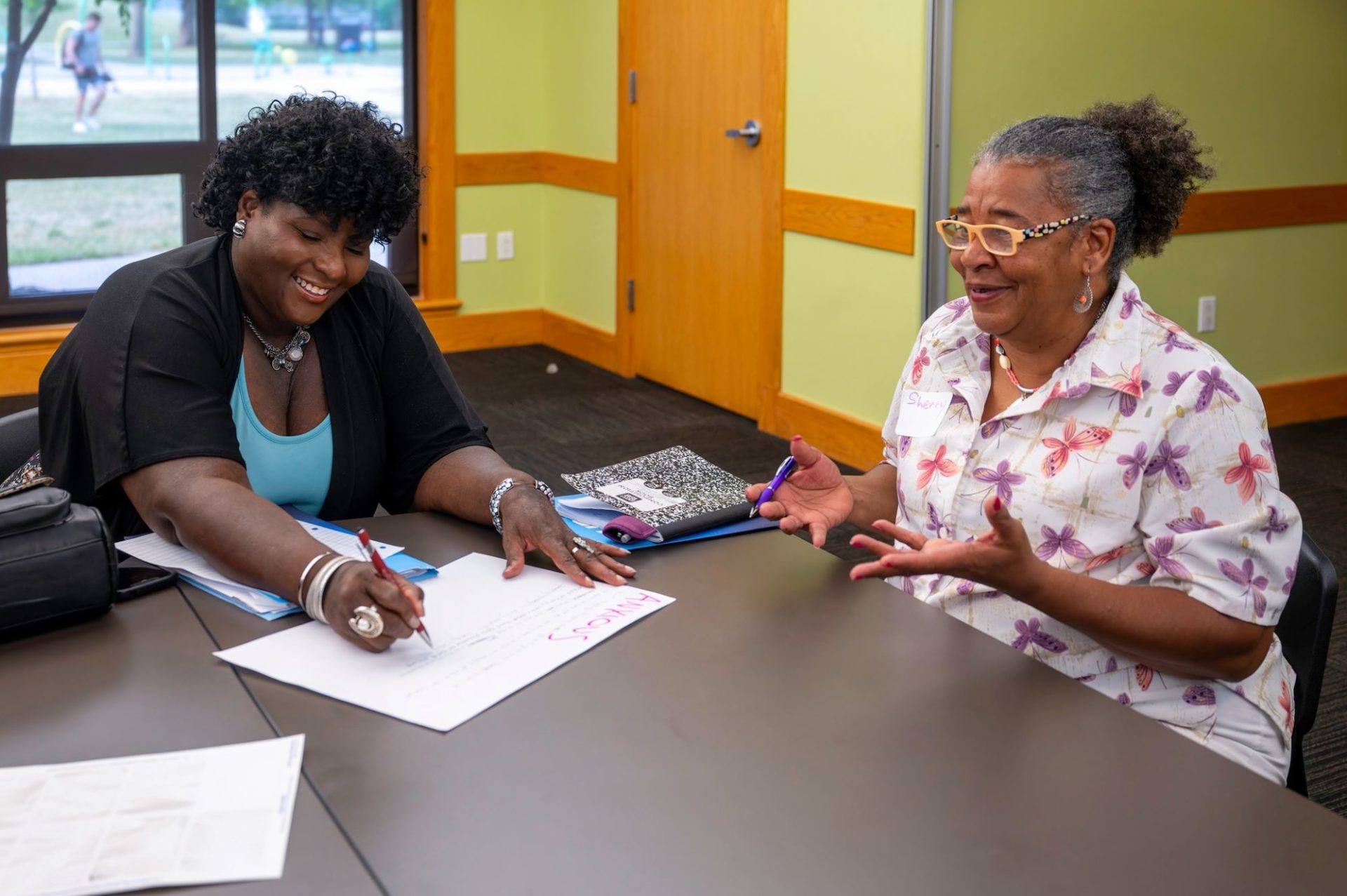 A younger Black woman and older Black woman are sitting across from one another at a table. The younger woman is writing on a large piece of paper.