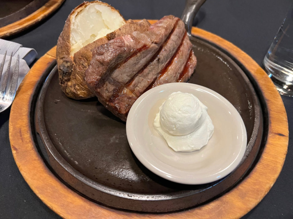 A ribeye leans against a baked potato beside a beige bowl of sour cream.