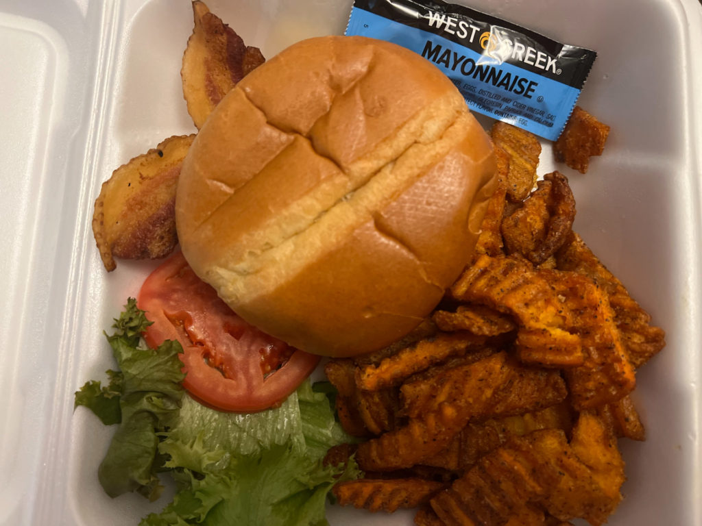 Half-pound burger with sweet potato fries in a styrofoam container with a blue packet of west creek mayonnaise.