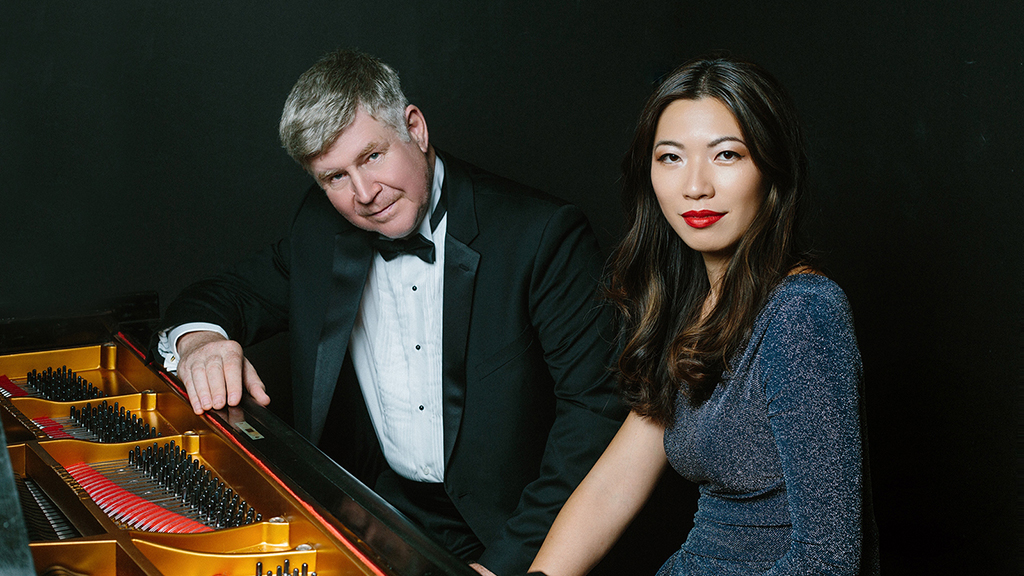 A man in a tuxedo sits next to a woman in a blue dress on a piano stool, while leaning on the piano. Both are looking into the camera.