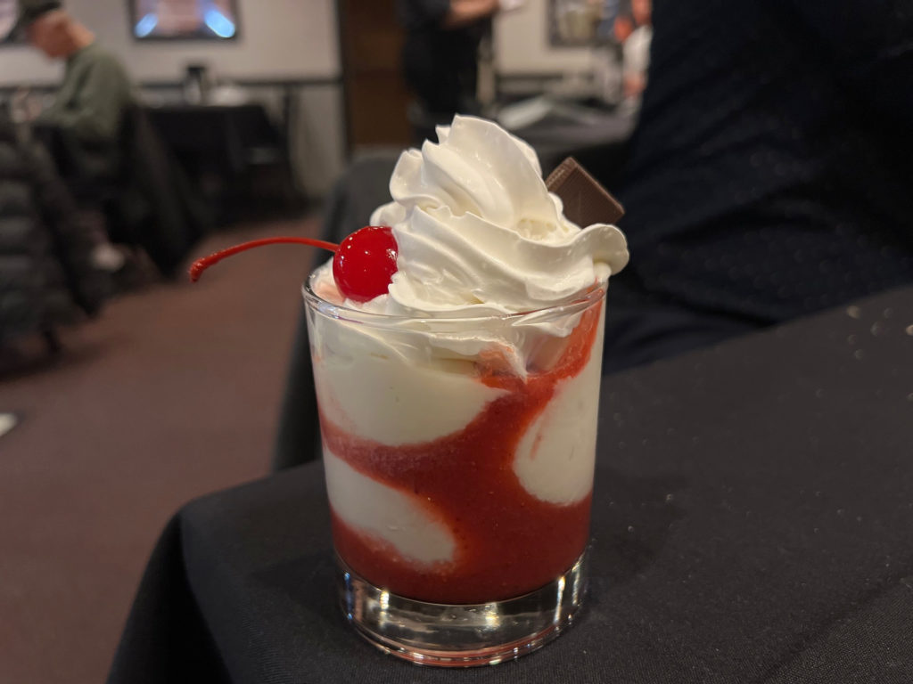 A glass cup of vanilla ice cream with whipped cream, a cherry, and a piece of chocolate.