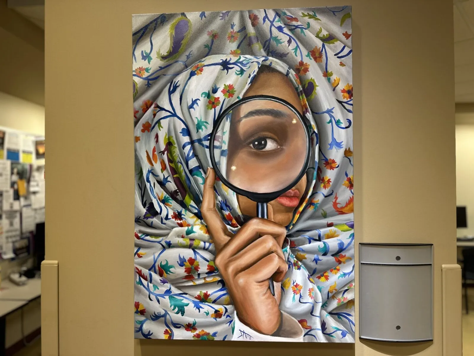 a vertical painting hangs on a tan wall; it features an upclose woman wearing a floral hijab holding a magnifying glass up to her eye