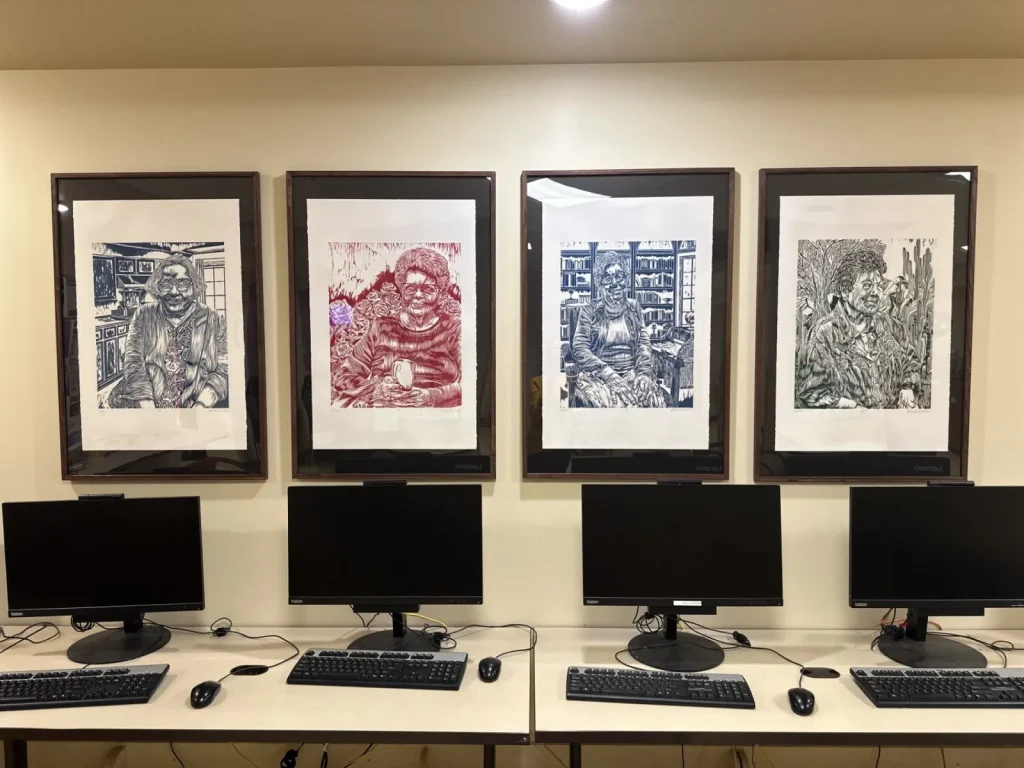 Four vertical artworks hang above computer stations. Each artwork is in a black frame with a white mat, and each features an outline of a person with a primary color: one is entirely in grey, another red, another blue, another grey. 