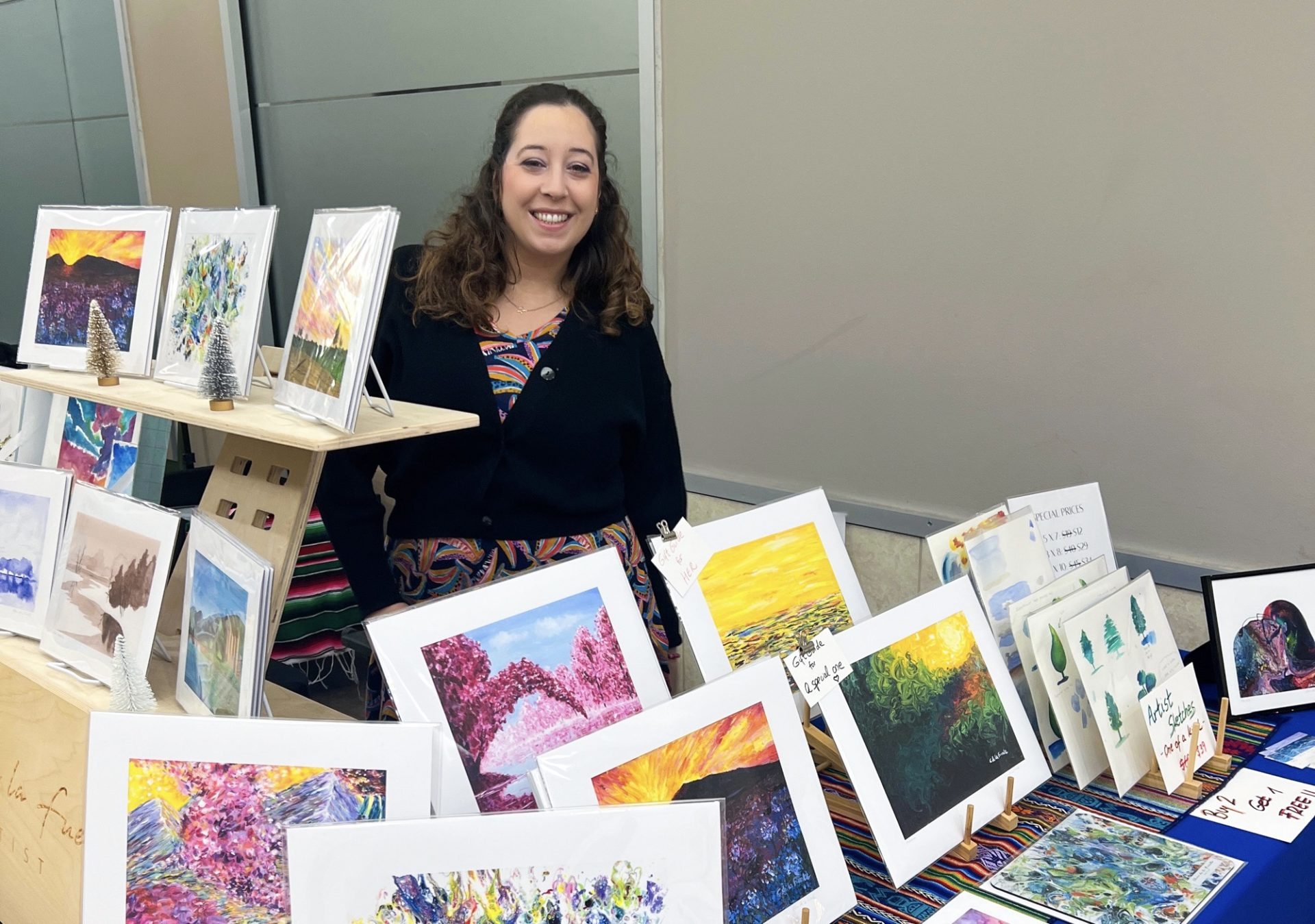 A woman in a black sweater and a colorful shirt stands behind a table of paintings