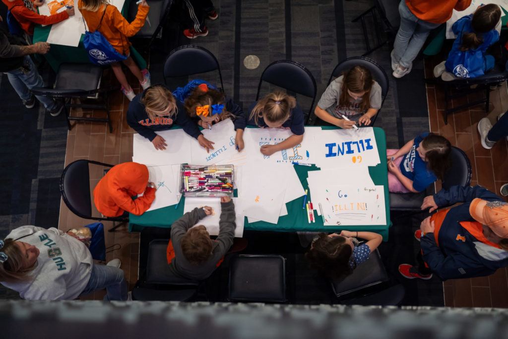 A bird's eye view of a table with eight children around it, making signs to cheer for the Illini women's basketball team.