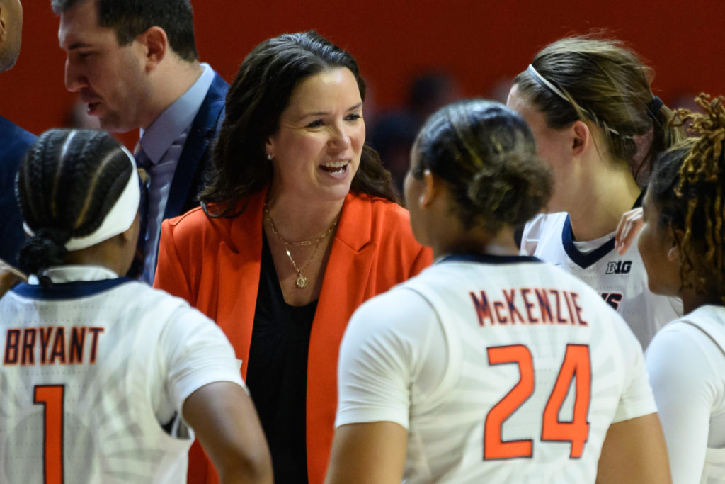 Illini coach Shauna Green is a white woman with long, dark hair. She is standing among Illini women's basketball players. Green is wearing an orange blazer.