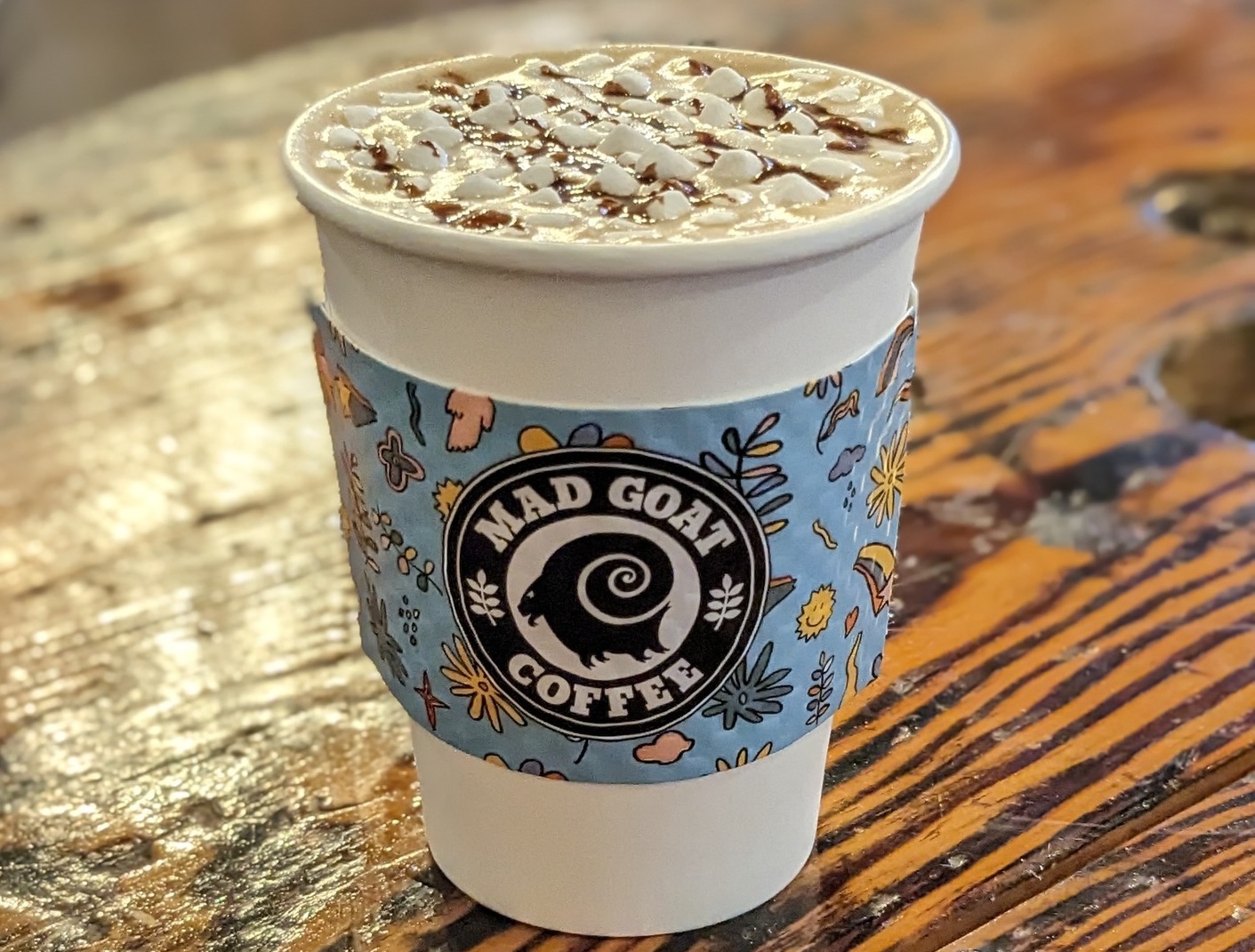 A white paper cup with a s'mores latte. There is a blue coffee cup holder with the Mad Goat Coffee logo on it.