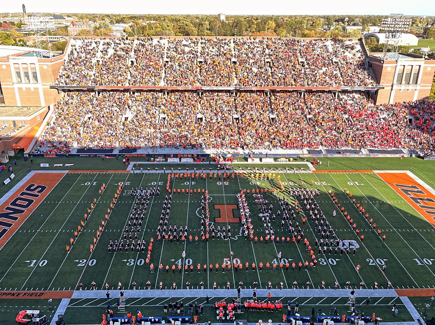 View of Zuppke Field at Memorial Stadium, University of Illinois. The Marching Illini band is in a block-I formation on the field. Most of the field is cast in shadow. The stands are full of spectators.