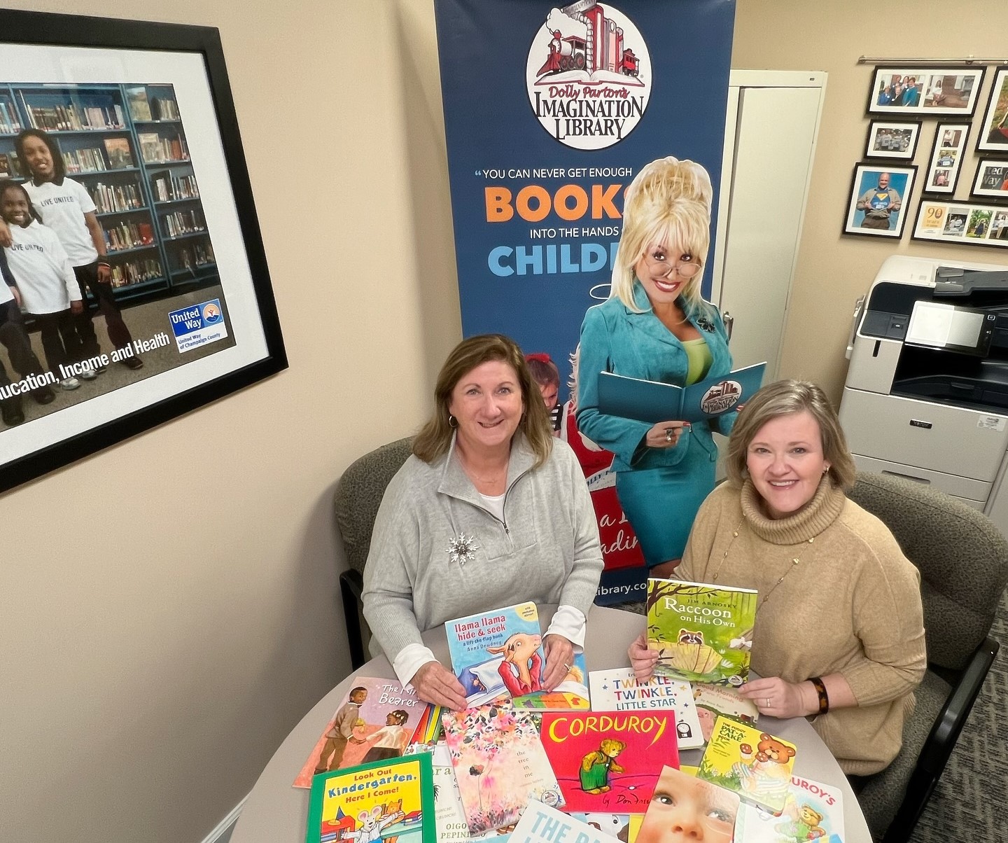 Two white women are seated at round table covered with colorful children's books. There is a standup banner behind them featuring an image of Dolly Parton.