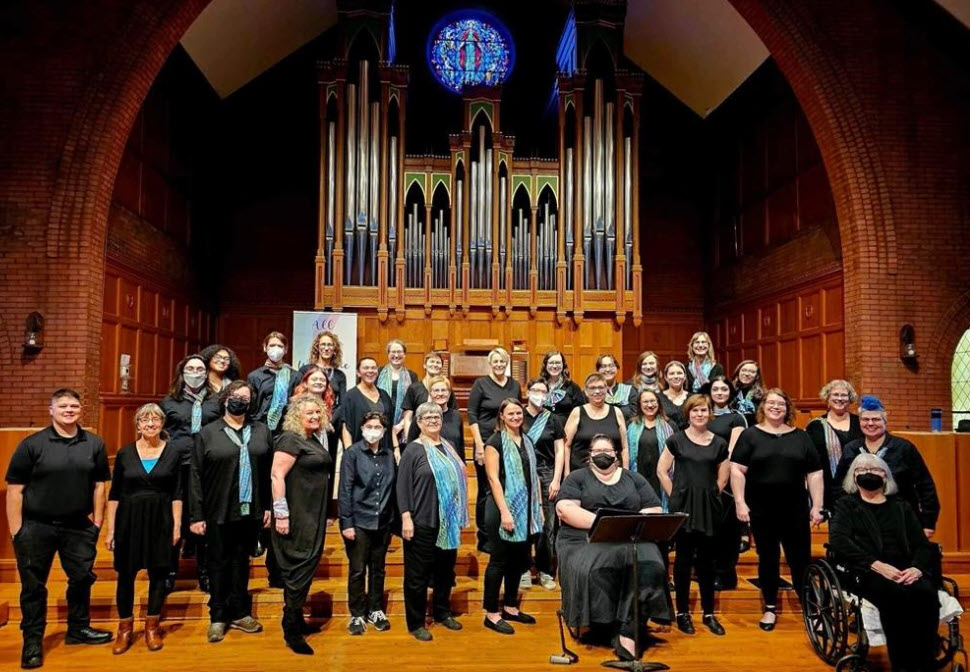 A gathering of individuals is seen on a stage, all dressed in black attire. The group is arranged in three rows, with most standing and two seated. The variety in their outfits includes plain shirts, scarves, and vests. One person is seated in a wheelchair. The backdrop is a grand pipe organ, set against brick-designed walls with a stained glass window above. The floor beneath them appears to be wooden. This scene takes place indoors. There are approximately 20 people present.