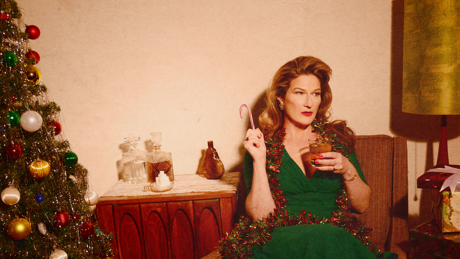 A woman in a green dress with a drink and a cigarette looking off to her left. There is a table with a red and white tablecloth, and the image gives off a holiday vibe.