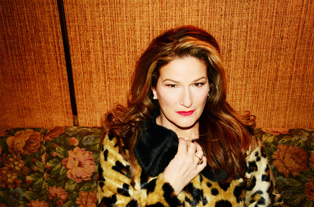 A woman sitting on a couch in a leopard print sweater. The couch has a floral design and the wall behind it is wood paneling.