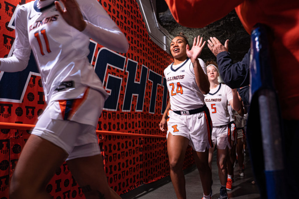 Illini women's basketball players Jada Peebles, Adalia McKenzie, and Gretchen Dolan are moving through an entrance tunnel toward a basketball court. They are wearing white Illinois uniforms.