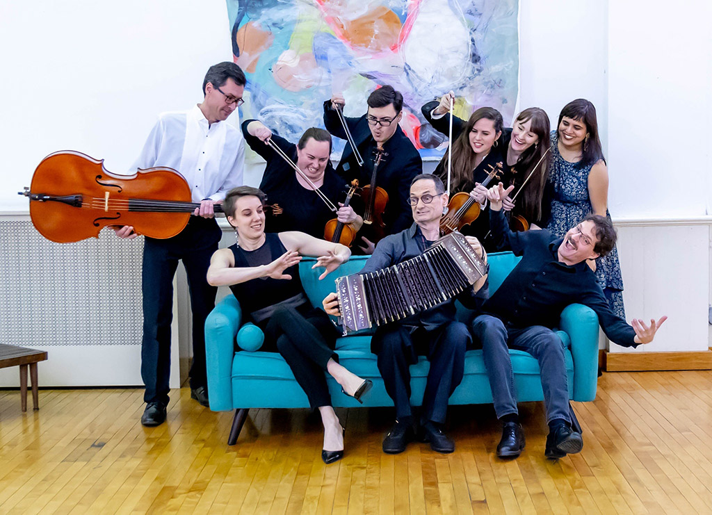 A group of 9 musicians, mostly string instrument players, smiling and jokingly posing as if they are going to attack their accordion player with their instruments. The accordion player is sitting on the couch playing his accordion defiantly.